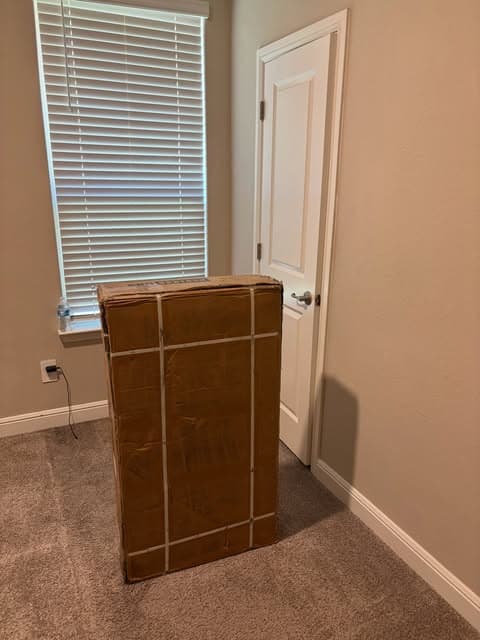 Large boxed flat-pack furniture item standing ready in a carpeted bedroom